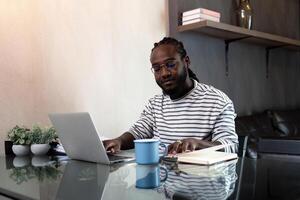 africano americano uomo Lavorando con il computer portatile computer a distanza mentre seduta a bicchiere tavolo nel vivente camera. nero tipo fare libero professionista opera a casa ufficio foto
