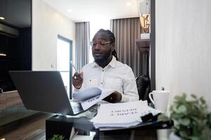 africano americano uomo Lavorando con il computer portatile computer a distanza mentre seduta a bicchiere tavolo nel vivente camera. nero tipo fare libero professionista opera a casa ufficio foto