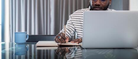 africano americano uomo Lavorando con il computer portatile computer a distanza mentre seduta a bicchiere tavolo nel vivente camera. nero tipo fare libero professionista mentre assunzione Appunti opera a casa ufficio foto