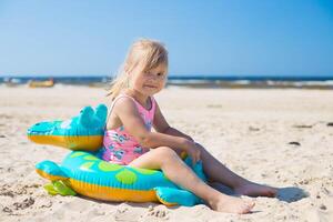 contento ragazza di europeo aspetto età di 5 seduta su un gonfiabile coccodrillo giocattolo a il spiaggia estate soleggiato giorno, guardare per il camera.family estate vocazione concetto. copia spazio. foto