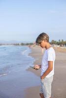 bello adolescente ragazzo nel bianca maglietta utilizzando smartphone a il spiaggia su estate vacanze, viaggio o viaggio nel Spagna .viaggi, vacanze, vacanze, la libertà concetto.lato Visualizza verticale . foto