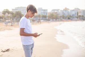 adolescente ragazzo utilizzando mobile Telefono a il spiaggia su estate vacanze, viaggio o viaggio nel Spagna .viaggio, vacanza, vacanze, la libertà concetto.lato Visualizza. foto