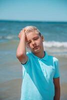 bello adolescente ragazzo di europeo aspetto con biondo capelli nel bianca corti, e un' blu maglietta sta nel il mare nel il acqua, e sembra per il telecamera. estate vocazione concetto.bello adolescente ragazzo ritratto concetto.verticale foto. foto