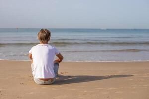 vicino su indietro Visualizza bello adolescente ragazzo nel bianca maglietta seduta a il spiaggia e godere un' estate vacanza guardare lontano, con spazio per testo.famiglia vacanze, vacanze, viaggio, viaggio concetto. foto