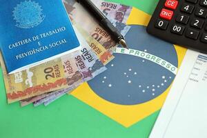 brasiliano opera carta e sociale sicurezza blu libro e reale i soldi fatture con calcolatrice e penna su bandiera di federativo repubblica di brasile foto