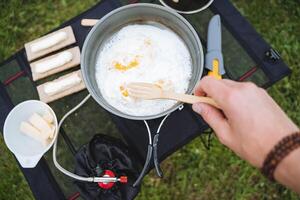 un' mano detiene un' forchetta fatto di Di legno, un' persona mangia strapazzate uova a partire dal un' frittura padella, campeggio cibo, prima colazione nel natura, campeggio nel il foresta, cucinando su un' gas bruciatore. foto