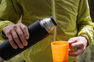 il thermos è tiro avvicinamento contro il sfondo di il umano corpo, mani Tenere un' tazza, un' bottiglia di termo con Tè, all'aperto ricreazione, escursioni a piedi vacanze, un' delizioso bere. foto