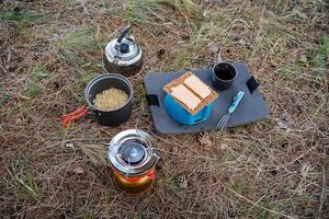 campeggio pasto prima colazione su il erba, formaggio panini, spaghetto pentola, gas bruciatore, metallo bollitore con Tè, boccale di Tè, campeggio pasti, cucinando foto