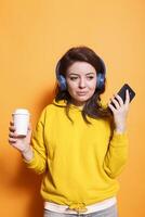 ritratto di femmina adulto afferrare mobile dispositivo e tazza di caffè mentre utilizzando cuffia. caucasico donna godendo musica su senza fili cuffia mentre utilizzando smartphone e avendo bere. foto