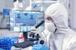 concentrato scienziato nel ppe attrezzatura guardare in il microscopio nel laboratorio. scienziato nel protettivo completo da uomo seduta a posto di lavoro utilizzando moderno medico tecnologia durante globale epidemico. foto