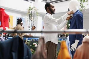 capi di abbigliamento memorizzare dipendente regolazione maschio completo da uomo su indossatrice mentre Lavorando nel shopping centro commerciale. moda boutique africano americano assistente fissaggio formale giacca su manichino modello foto