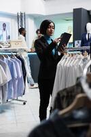 capi di abbigliamento memorizzare asiatico donna lavoratore maschiatura su digitale tavoletta touch screen mentre Lavorando. shopping centro boutique dipendente utilizzando merce inventario Software App su portatile aggeggio foto