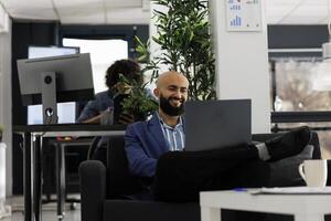 sorridente arabo uomo d'affari avendo conferenza con collaboratore nel un' aziendale ufficio. progetto manager utilizzando un' il computer portatile per telelavoro incontro nel inizio su azienda coworking spazio foto
