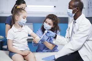 africano americano medico controlli e ossequi il ferita di un' caucasico ragazza nel un' clinica. maschio medico con un' laboratorio cappotto è Assistito di un' femmina infermiera nel blu scrub con mettendo un' garza su il paziente. foto