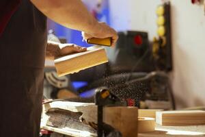 craftsperson a banco di lavoro utilizzando Manuale carta vetrata per levigatrice legname bloccare, assemblaggio mobilia nel la lavorazione del legno negozio. falegname levigante pezzo di Di legno, godendo Fai da te passatempo, vicino su foto