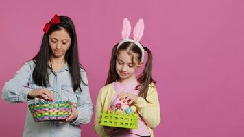 gioioso fiducioso bambino e madre mostrando Pasqua cesti su telecamera, decorazione festivo accordi per primavera vacanza. contento scolara con coniglietto orecchie in posa con sua mamma nel studio. telecamera un. foto