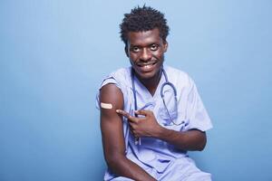 africano americano medico infermiera avendo adesivo bendare dopo vaccinazione contro coronavirus. vaccinati maschio infermiera con scrub puntamento a vaccino tiro toppa per immunità. foto