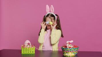 entusiasta giovane ragazza giocando sbirciare un' fischio nel davanti di telecamera, utilizzando dipinto Pasqua uova contro rosa sfondo. gioioso bello bambino piccolo sensazione eccitato di primavera vacanza festa. telecamera un. foto