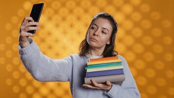 sorridente donna Tenere mucchio di libri, godendo lettura passatempo, assunzione selfie con cellulare. allegro signora con pila di romanzi nel braccia fare immagini con mobile Telefono, studio sfondo, telecamera B foto