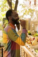 ritratto di sorridente maschio individuale degustazione Mela prima acquisto bio produrre, shopping per naturale biologico prodotti a agricoltori mercato. giovane nero uomo godendo cibo degustazione fresco Locale produrre. foto