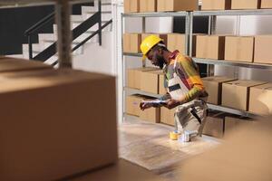 magazzino supervisore analizzando merce lista di controllo, in piedi accanto cartone scatole nel Conservazione camera. africano americano manager indossare casco e industriale complessivamente Lavorando nel magazzino foto