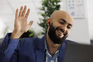 sorridente imprenditore frequentando a distanza conferenza chiamata e saluto dipendenti. arabo esecutivo manager agitando Ciao per squadra durante virtuale conferenza per discutere progetto foto