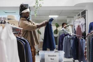 africano americano uomo mettendo casuale camicia nel donazione scatola mentre shopping nel boutique. centro commerciale cliente lancio via secondo mano abbigliamento nel contenitore per caritatevole organizzazione foto