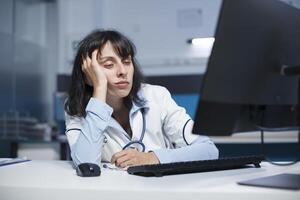 logoro su femmina medico seduta a un' scrivania con un' del desktop pc dopo un' lungo giorno nel Ospedale. caucasico donna con sua mano per sua viso, guardare stanco mentre fare assistenza sanitaria ricerca. avvicinamento sparo. foto