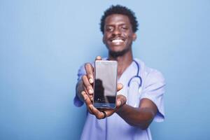 maschio infermiera guardare a telecamera mentre trasporto cellula Telefono con vuoto schermo, contro blu sfondo. dimostrando medico competenza, nero uomo indossa scrub e stetoscopio mentre Tenere mobile dispositivo. foto