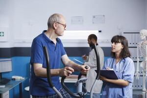 anziano paziente è visto ricevente fisioterapia su stazionario bicicletta, Assistito di femmina infermiera vestito nel blu scrub. anziano uomo è ottenere riabilitativo cura con Aiuto a partire dal Salute professionale. foto