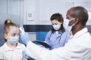 donna nel blu scrub scrittura Appunti mentre nero uomo nel un' laboratorio cappotto controlli temperatura di caucasico bambino. Immagine Spettacoli maschio medico l'esame paziente con assistenza a partire dal infermiera. foto