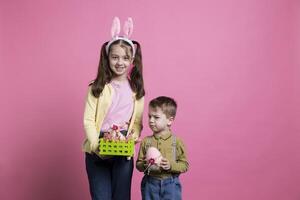 giocoso bambini mostrando dipinto uova e decorazione per Pasqua celebrazione festa, in piedi insieme al di sopra di rosa sfondo. fratello e sorella sensazione allegro e entusiasta di aprile evento. foto