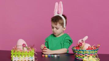 adorabile poco giovanotto crea fatto a mano ornamenti per Pasqua Domenica e pittura uova con francobolli e spazzole. lieta giovane ragazzo utilizzando arte materiale per colore e decorare. telecamera un. foto