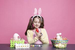 carino poco ragazza colorazione uova con acquerello e pennello per Pasqua preparativi, decorazione festivo ornamenti contro rosa sfondo. piccolo bambino piccolo avendo divertimento con decorazioni. foto