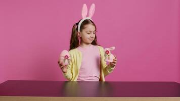 piccolo allegro ragazza mostrando colorato decorato ornamenti su telecamera, la creazione di fatto a mano festivo disposizioni. giovane bambino sensazione eccitato di Pasqua domenica, detiene coniglio e uovo. telecamera b. foto