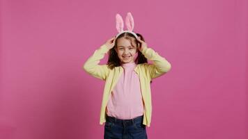 adorabile carino bambino mettendo coniglietto orecchie e agitando a telecamera, godendo Pasqua Domenica celebrazione contro rosa sfondo. sorridente allegro scolara con trecce detto ciao. telecamera b. foto