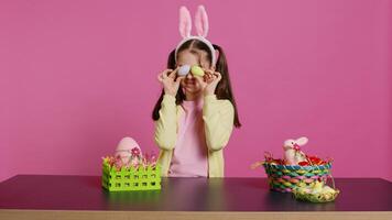 entusiasta giovane ragazza giocando sbirciare un' fischio nel davanti di telecamera, utilizzando dipinto Pasqua uova contro rosa sfondo. gioioso bello bambino piccolo sensazione eccitato di primavera vacanza festa. telecamera b. foto