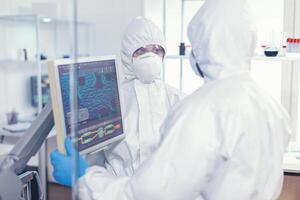 collaboratori nel tuta discutere di virus Evoluzione vestito nel ppe completo da uomo nel moderno assistenza sanitaria facilità. medici l'esame virus Evoluzione utilizzando alto Tech ricercando diagnosi contro covid19 foto