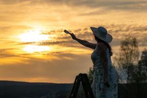 un' donna nel un' vestito sta su un' di legno scala e vernici il tramonto foto