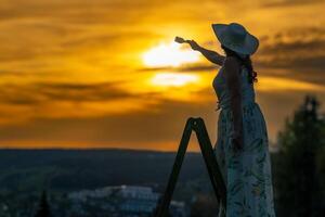 un' donna nel un' vestito sta su un' di legno scala e vernici il tramonto foto