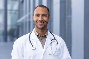 ritratto di riuscito e sorridente africano americano medico, uomo nel medico cappotto con stetoscopio guardare a telecamera e sorridente, al di fuori moderno ospedale foto