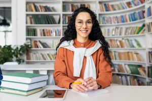 contemporaneo in linea precettore seduta a tavolo nel biblioteca circondato di scaffali pieno con libri. bella donna indossare un arancia giacca con bianca felpa con cappuccio Tenere giallo penna a personale posto di lavoro. foto