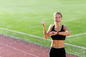 sportivo giovane donna atleta in piedi nel il stadio indossare cuffie e sorridente a il telecamera, controllo il tempo su il fitness braccialetto, mostrando un' vittoria gesto con sua mano. foto