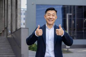 ritratto di un' giovane riuscito asiatico maschio uomo d'affari in piedi al di fuori un ufficio edificio nel un' completo da uomo, sorridente, guardare a il telecamera e mostrando il super cartello con il suo dita. foto