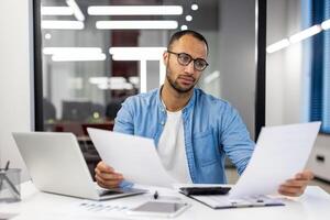 foto di un' grave giovane uomo contabile e finanziario analista seduta nel il ufficio a il scrivania e Lavorando con documenti.