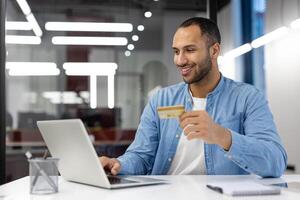 un' uomo è sorridente mentre Tenere un' credito carta nel davanti di un' il computer portatile. lui è probabile utilizzando il carta per rendere un' Acquista o dai un'occhiata il suo equilibrio. concetto di convenienza e alleviare foto