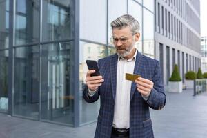 insoddisfatto anziano uomo nel completo da uomo Tenere carta e Telefono nel mani, in piedi su strada vicino ufficio, senza esito in linea pagamento. foto