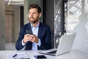 ritratto di un' riuscito giovane uomo uomo d'affari, proprietario seduta a un' scrivania nel il ufficio nel un' completo da uomo, Tenere un' mobile Telefono e guardare con fiducia per il lato. foto