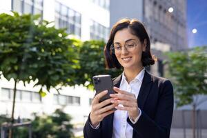 professionale donna d'affari nel bicchieri utilizzando sua smartphone con un' Sorridi, in piedi all'aperto con ufficio edifici e verdura nel il sfondo. foto