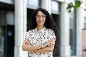 professionale e fiducioso ispanico donna con Riccio capelli, indossare bicchieri e un' polka punto camicia, sorrisi calorosamente nel un' moderno città ambiente. foto